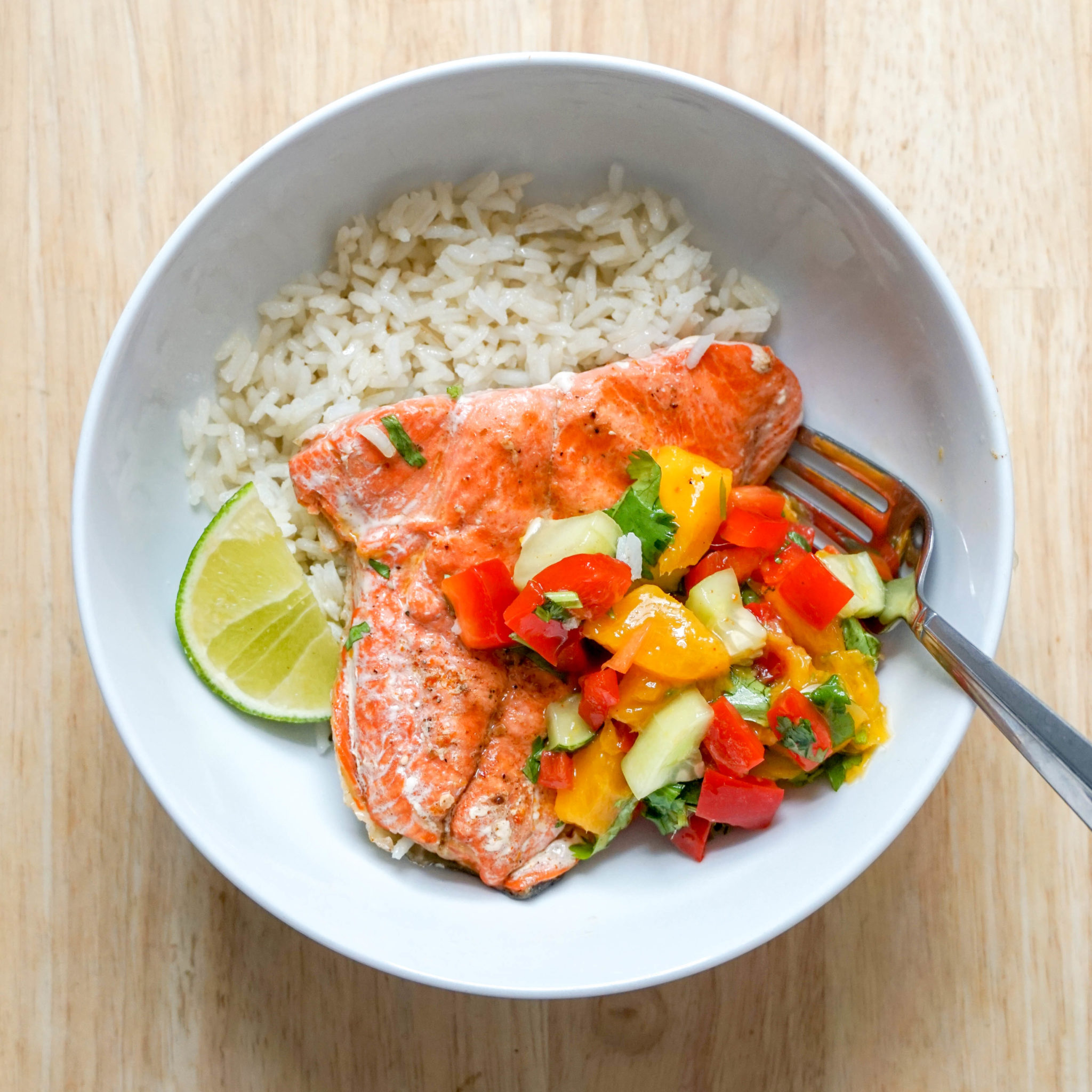 White round bowl filled with pink pan-seared salmon and colorful spicy mango salsa over rice. Served with a lime wedge 