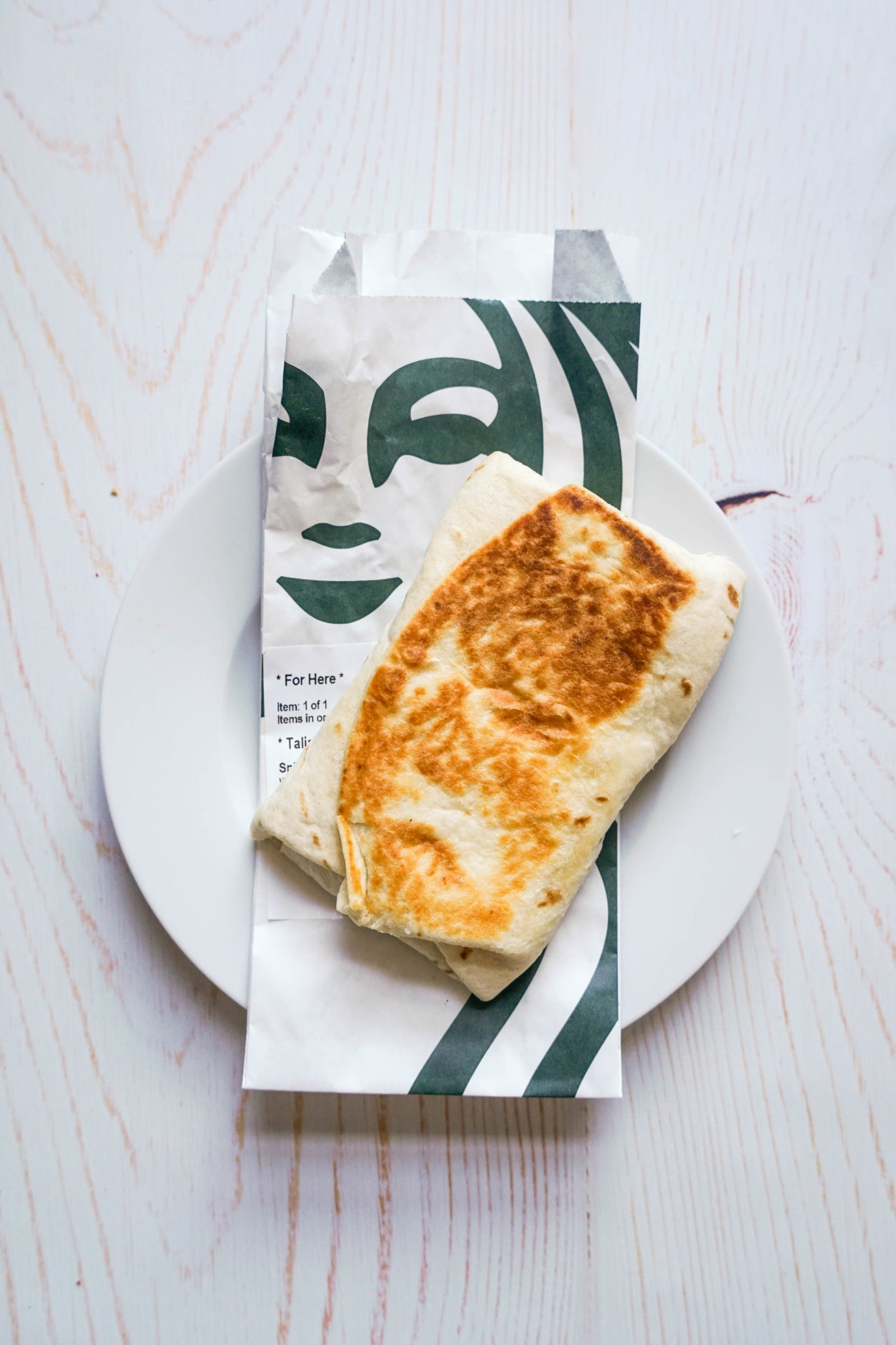 There is a golden breakfast wrap laying on top of a Starbucks wrapper, that has the company logo in green. Both are on top of a white plate