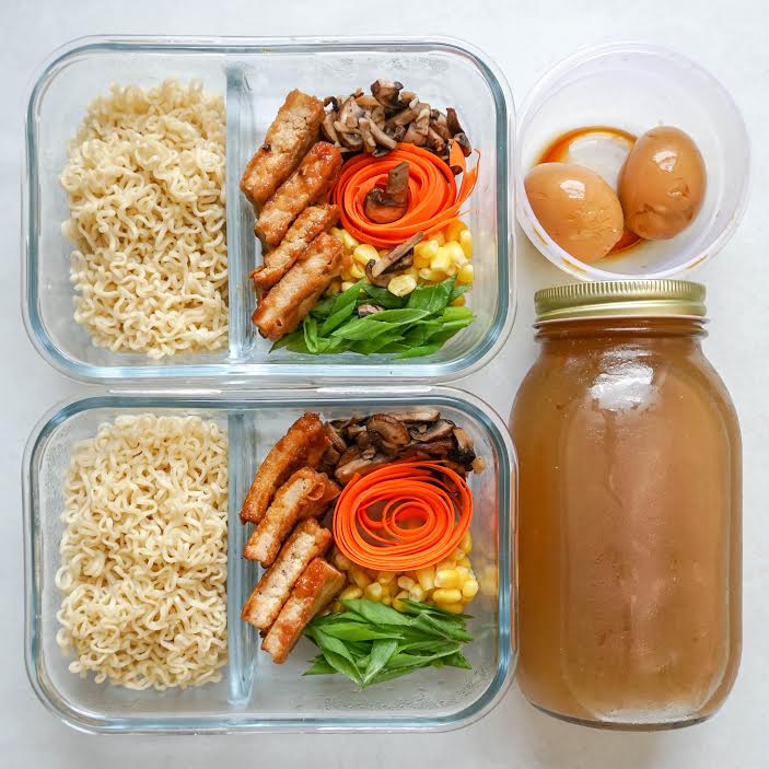 Two rectangular containers with dividers separating cooked ramen noodles from tofu, mushrooms, carrots, corn, and snap peas. There is a mason jar full of dark brown broth and a small round container with two marinated eggs.