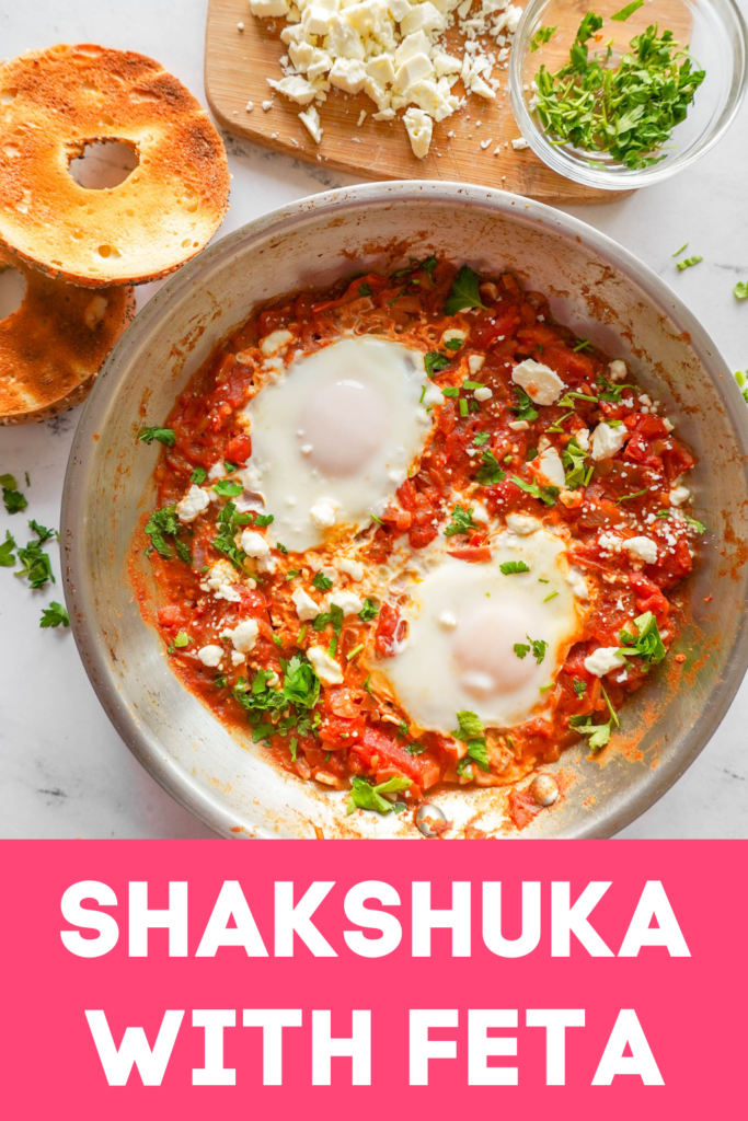 stainless steel pan with red tomato mixture and two cooked eggs with bits of green parsley and feta on top (shakshuka). Bagel in the background