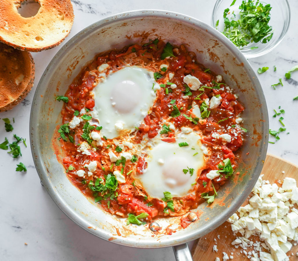 stainless steel pan with red tomato mixture and two cooked eggs with bits of green parsley and feta on top (shakshuka). Bagel in the background