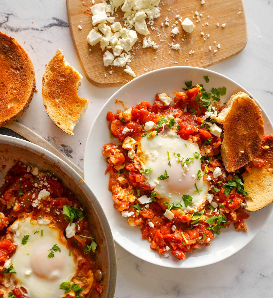 A plate of shakshuka- red tomato mixture and a cooked egg with feta and parsley on top. Served with bread. Stainless steel pan on the side.