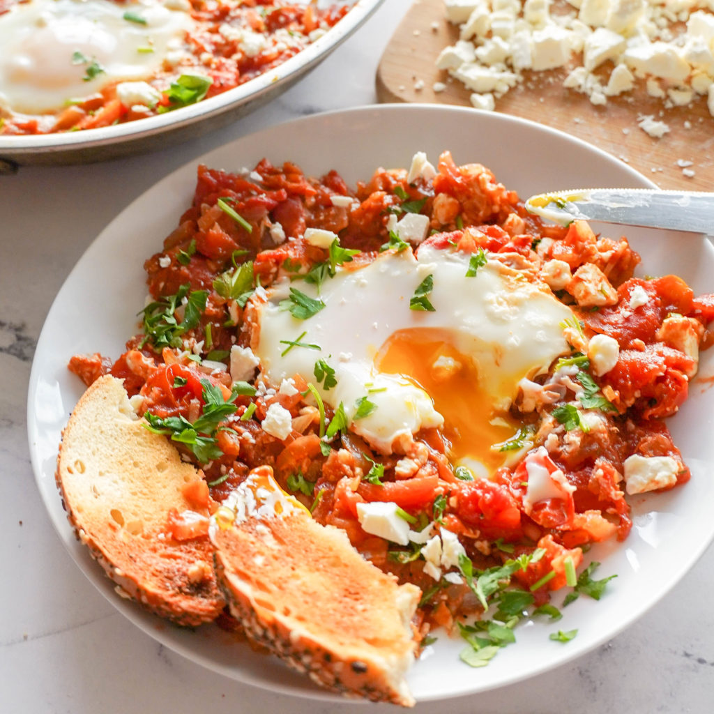 Close up of Shakshuka- red tomato mixture with a cooked egg on top, the egg is broken and the yolk in dripping out. Topped with feta and parsley and served with bread.