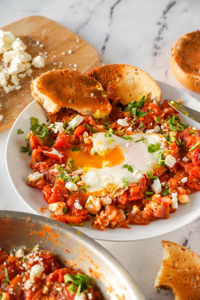Close up of Shakshuka- red tomato mixture with a cooked egg on top, the egg is broken and the yolk in dripping out. Topped with feta and parsley and served with bread.