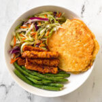 A round bowl is filled with corn fritters, green beans, tempeh, and coleslaw