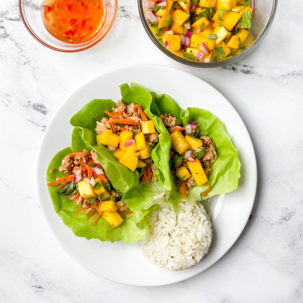 three open chicken lettuce wraps next to a scoop of rice on a white place with mango salsa and chili dipping sauce in the background.