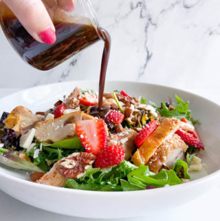 bowl full of strawberry, leek and chicken salad with a hand pouring balsamic dressing out of a small glass container on top of the salad.