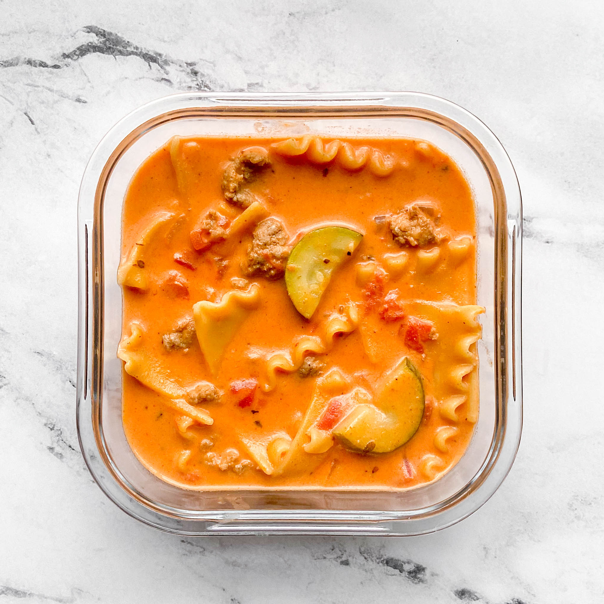 a square container of lasagna soup on a white and grey background. The soup has noodles, zucchini, meat, and an orange broth