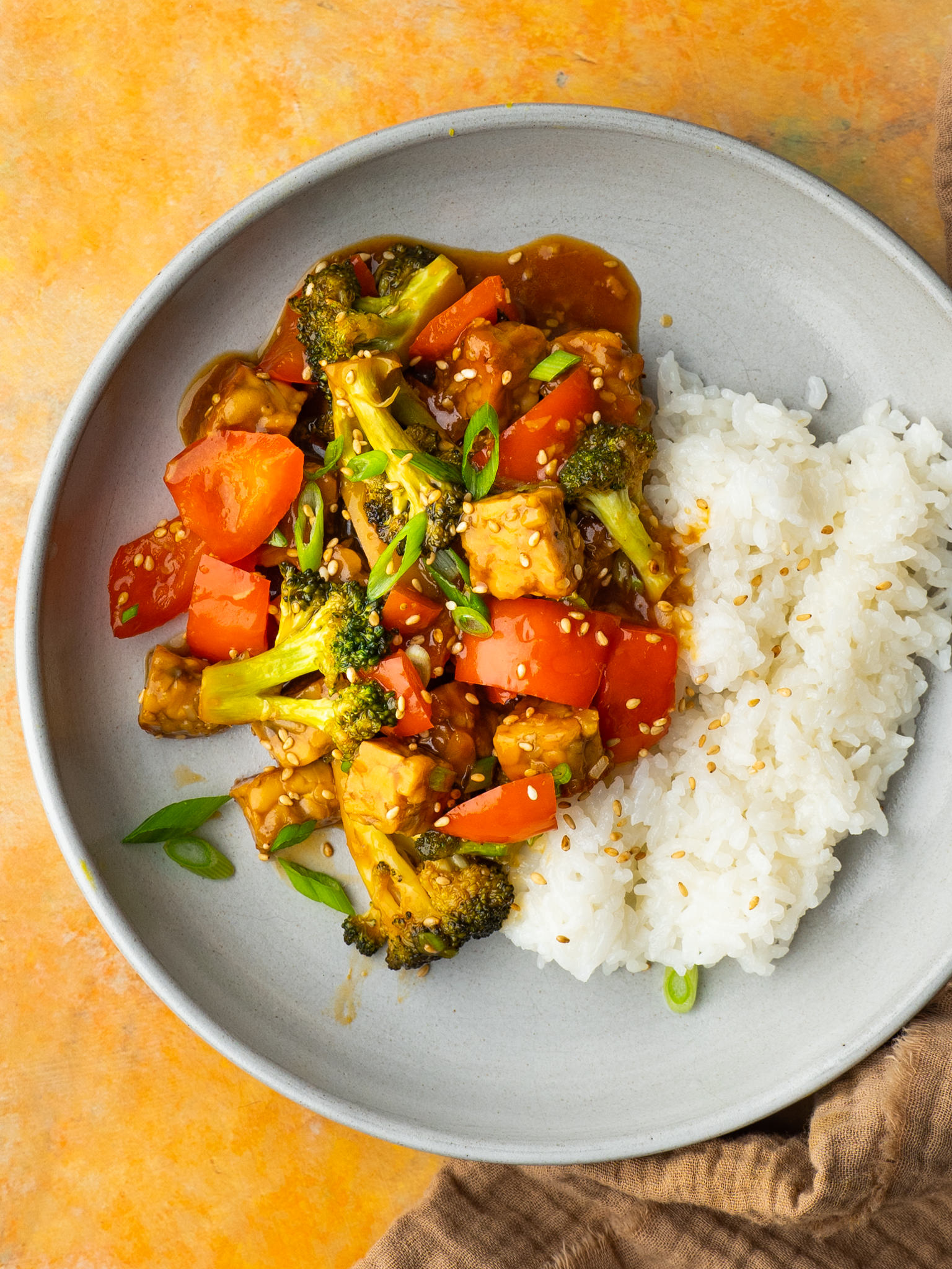A stir fry with broccoli and peppers is on a plate with a scoop of white rice next to it. The dish is garnished with sesame seeds.