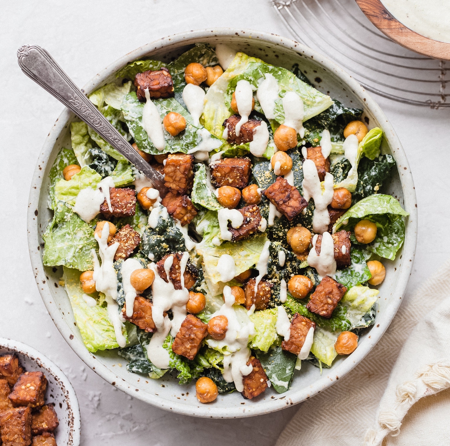 A green Caesar salad is in a white bowl. It is topped with chickpeas and a white Caesar dressing. There is a fork sticking out of the salad.
