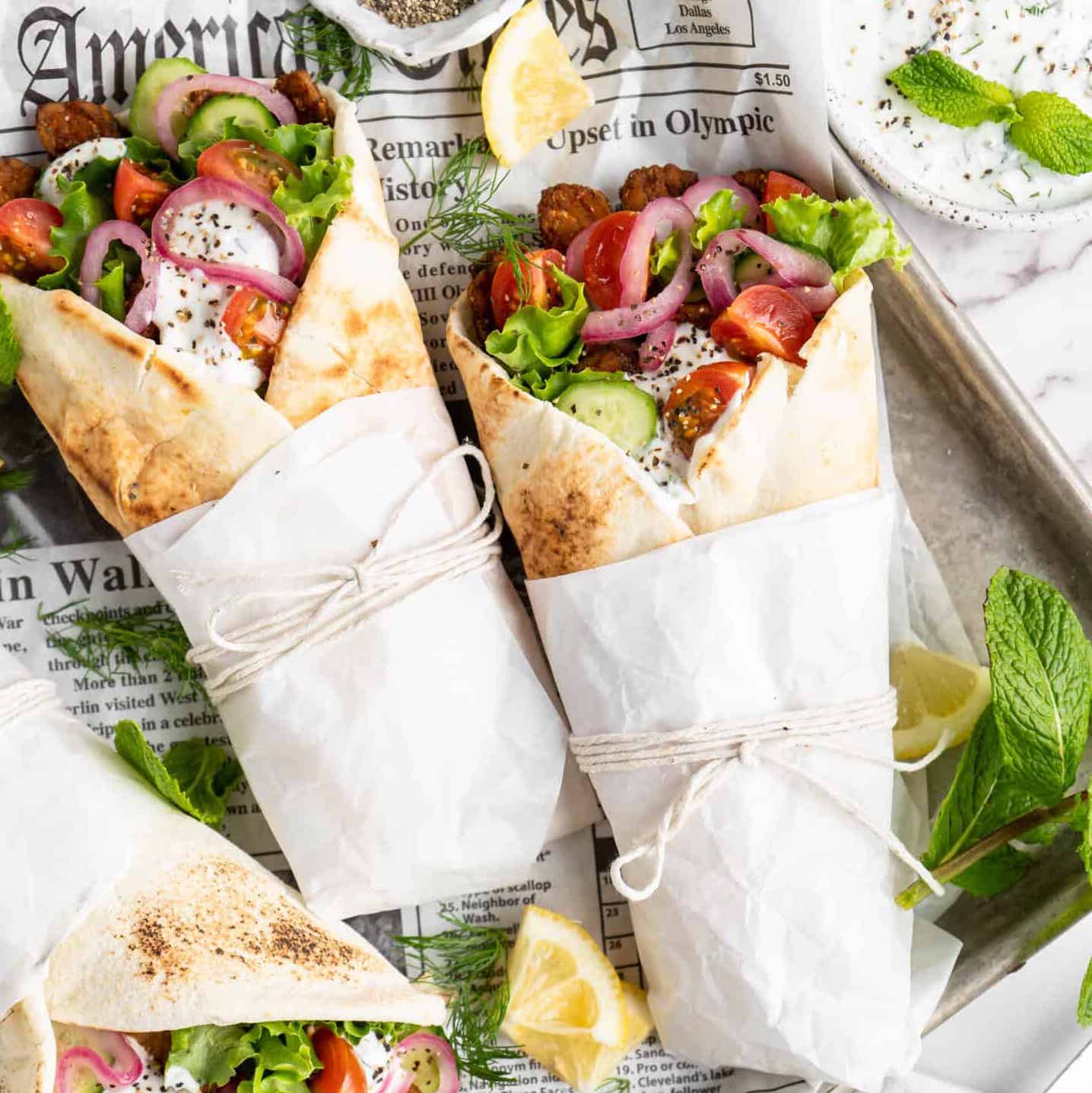 Two wrapped gyros are on a newspaper background. They have pickled red onions, cucumbers, tomatoes, and a white sauce.