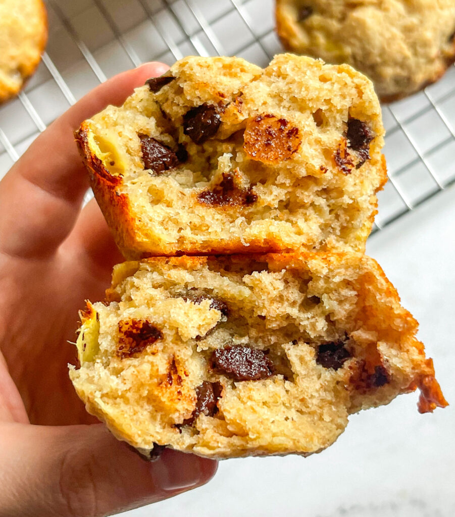 a banana muffin cut in half showing the inside with chocolate chips held between two fingers over a white marble background.