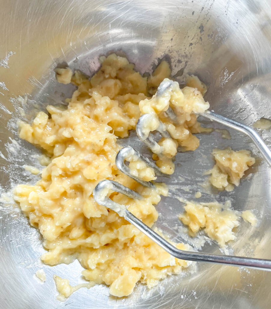 a close up shot of mashed banana in a stainless steel bowl with a mashing tool.