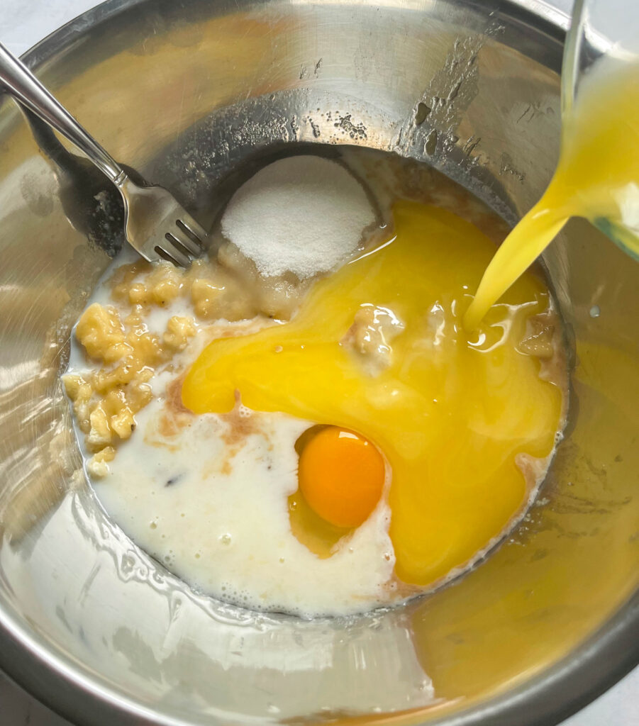 a stainless steel bowl filled with yellow melted butter, an egg, milk, sugar and mashed banana. A fork is sticking out of the bowl.