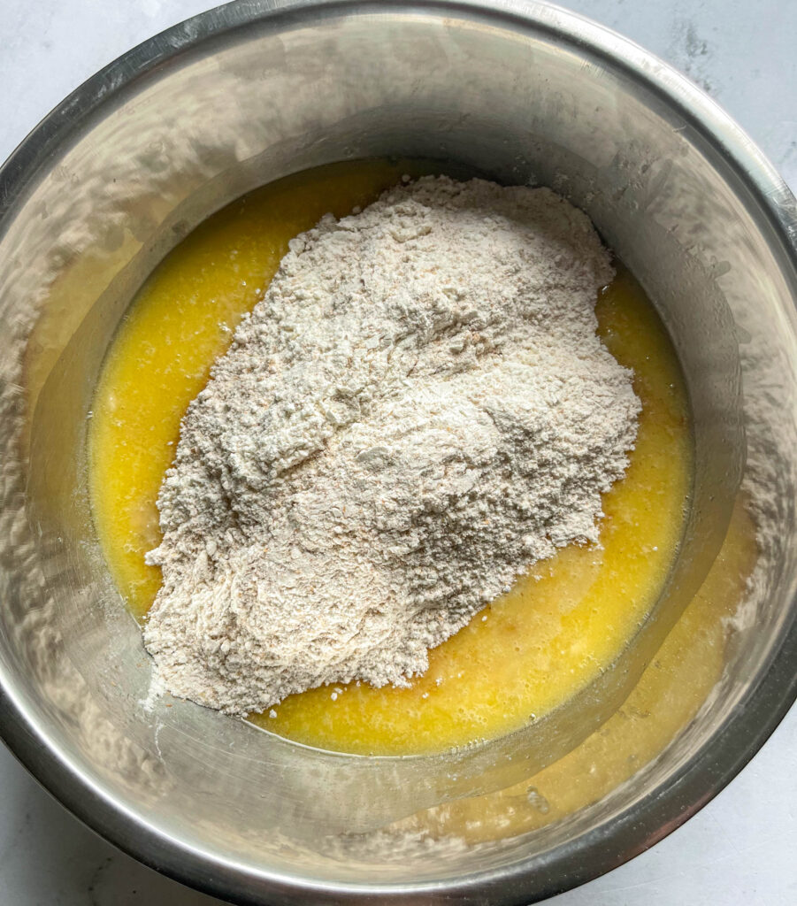 a stainless steel bowl filled with the wet banana muffin batter ingredients and the dry ingredients such as flour poured on top.