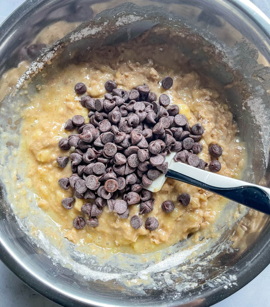 a silver stainless steel bowl filled with banana muffin batter. there's a pile of chocolate chips sitting on top of the batter and a spatula sticking out of the bowl.