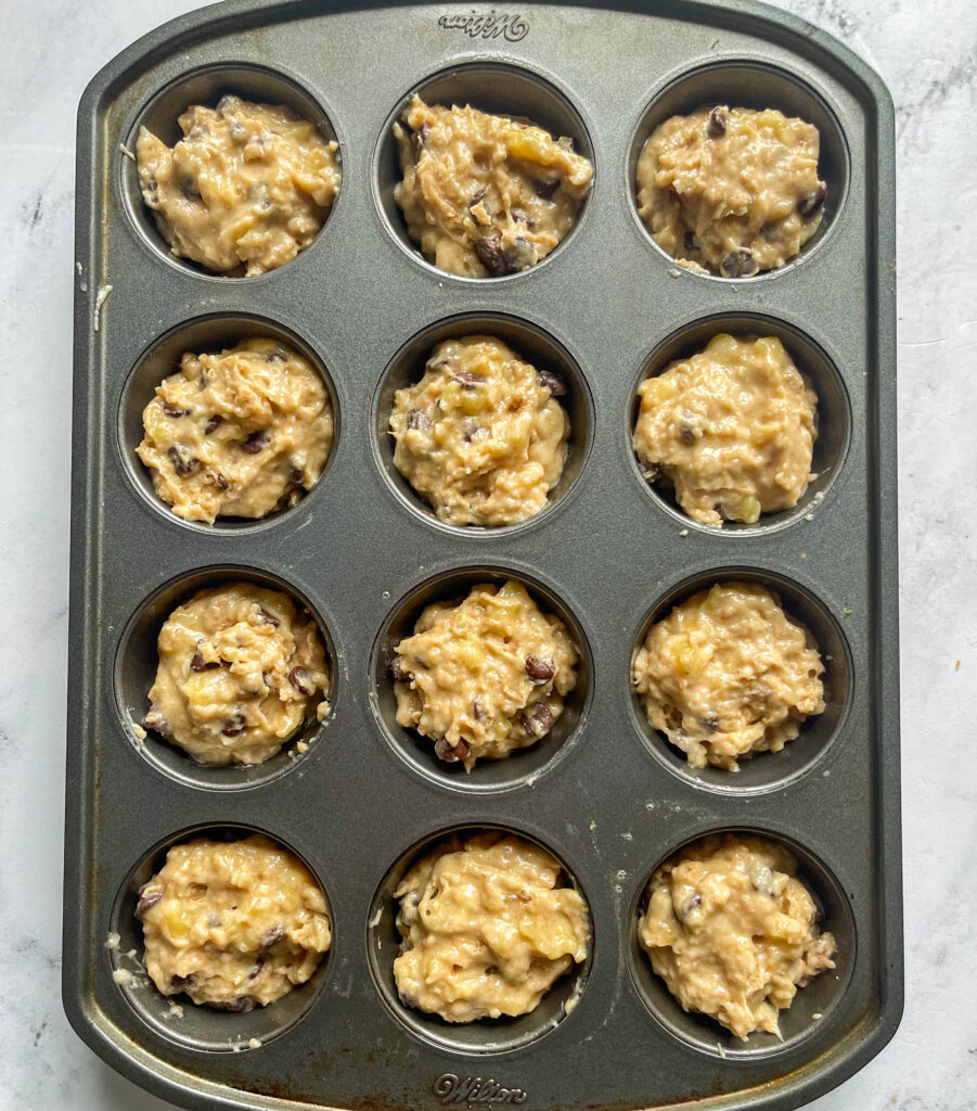 a muffin tin with 12 wells filled with banana chocolate chip muffin batter on a white marble background.