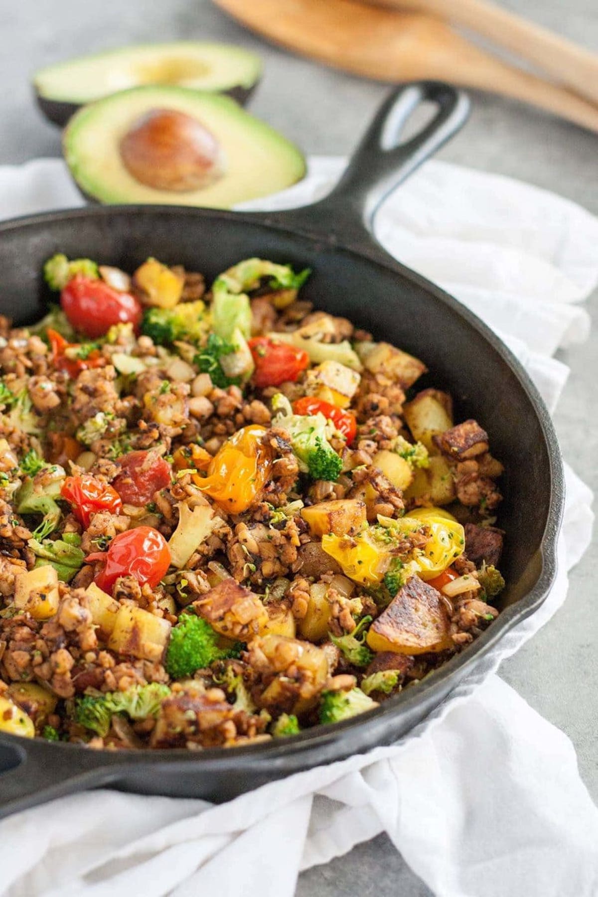 A cast iron skillet is filled with colorful tomatoes, broccoli, potatoes and tempeh