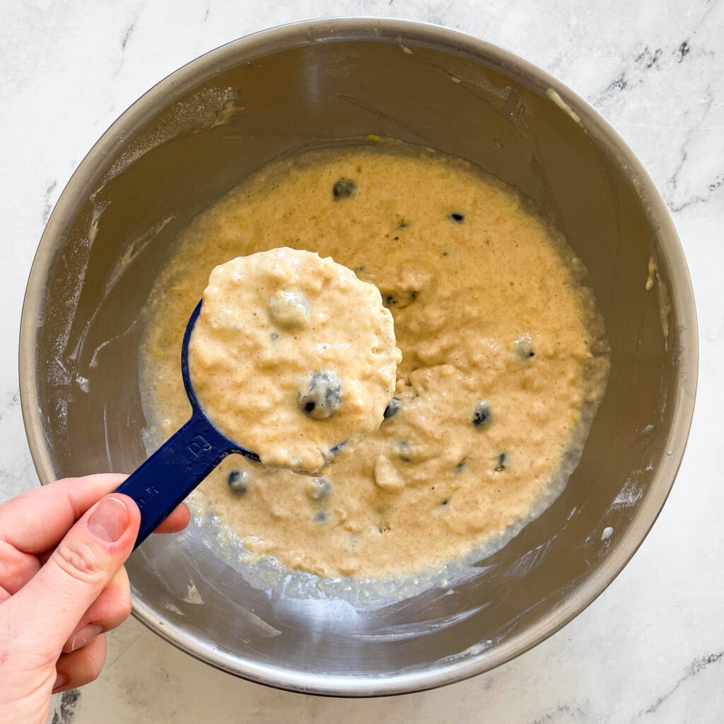 a bowl full of blueberry lemon pancake batter with a hand holding a scoop of batter in a measuring cup