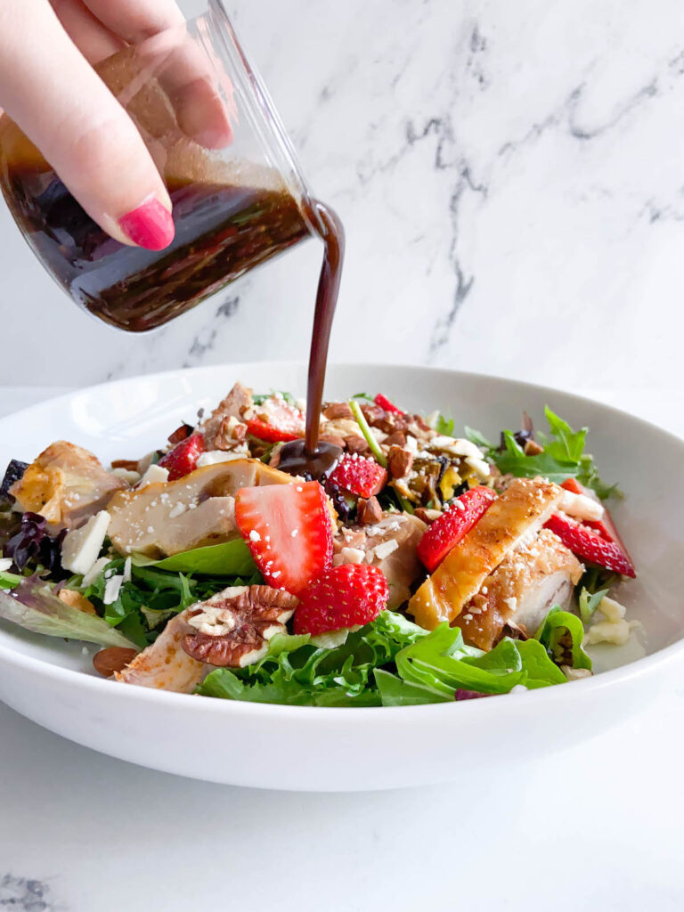 bowl full of salad with leeks, chicken and strawberries. a hand pouring balsamic dressing out of a small glass container over the bowl.